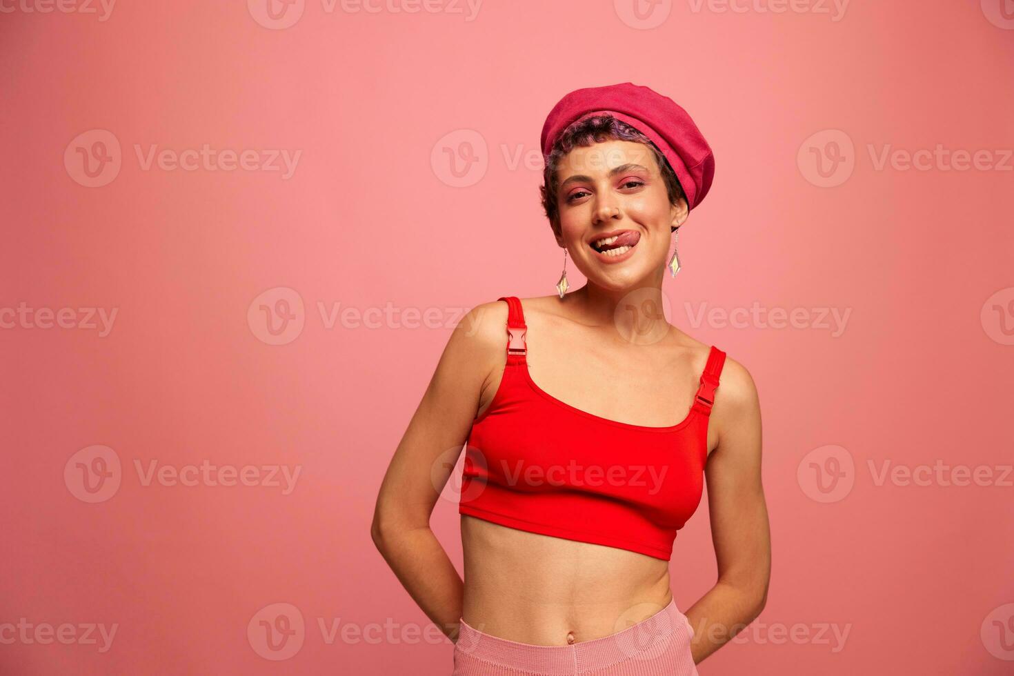 giovane atletico donna con un' corto taglio di capelli e viola capelli nel un' rosso superiore e un' rosa cappello con un atletico figura sorrisi e smorfie guardare a il telecamera su un' rosa sfondo foto