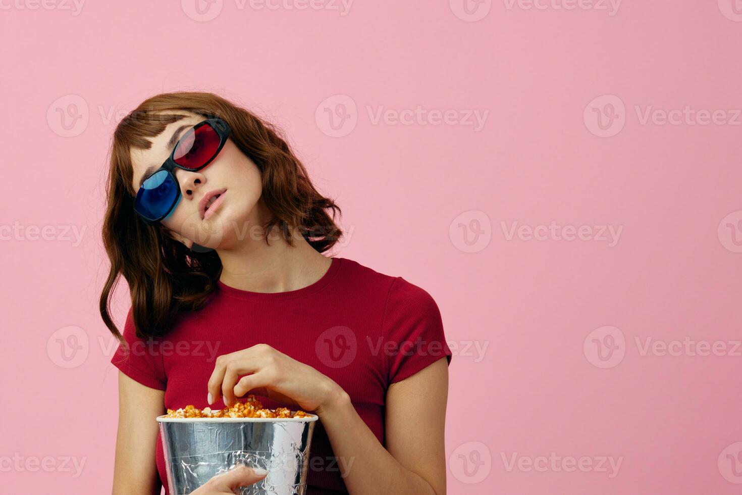 confuso concentrato carino testa Rossa signora nel rosso maglietta 3d bicchieri mangiare Popcorn orologio film in posa isolato su al di sopra di rosa studio sfondo. copia spazio striscione. moda cinema concetto. divertimento offrire foto