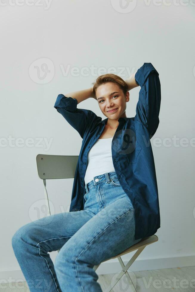 un' giovane donna seduta nel un' sedia a casa sorridente con denti con un' corto taglio di capelli nel jeans e un' denim camicia su un' bianca sfondo. ragazza naturale pose con no filtri foto