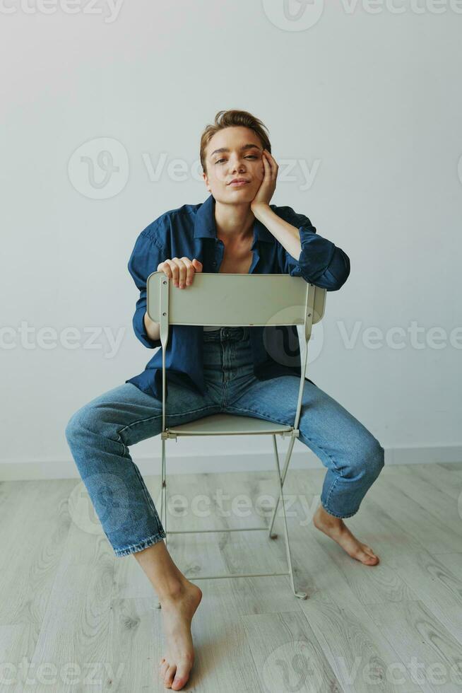 un' giovane donna seduta nel un' sedia a casa sorridente con denti con un' corto taglio di capelli nel jeans e un' denim camicia su un' bianca sfondo. ragazza naturale pose con no filtri foto