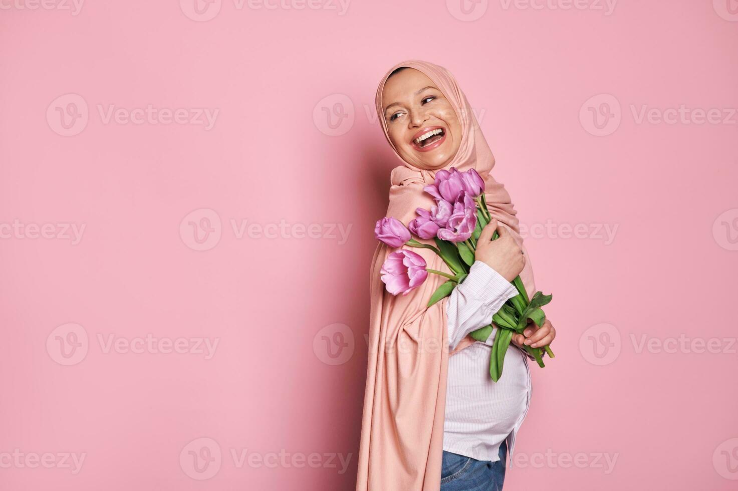 incinta arabo musulmano donna nel hijab detiene un' mazzo di tulipani per La madre di giorno, sorrisi guardare a parte, rosa sfondo foto