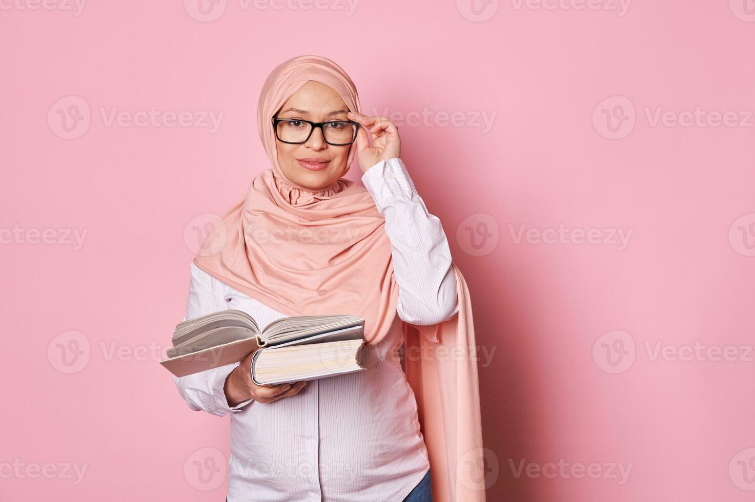 fiducioso arabo musulmano femmina educatore affascinante incinta donna nel rosa hijab e occhiali, Tenere prenotare, guardare a telecamera foto