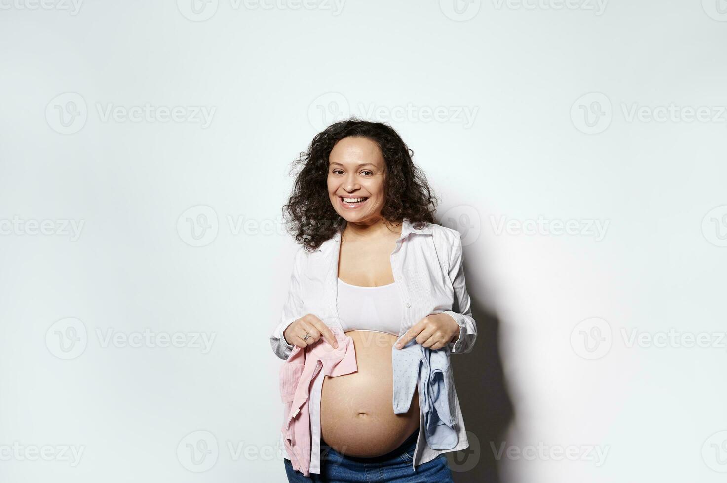 sorridente incinta donna Tenere blu e rosa neonato body vicino sua gonfiarsi. aspettandosi per un' bambino, ragazzo o ragazza, Gemelli foto