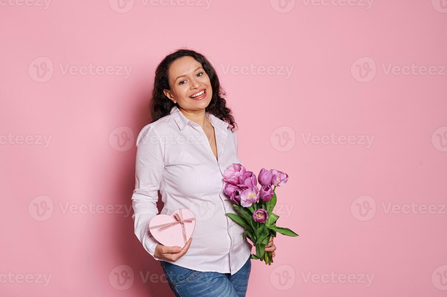 sorridente incinta donna, esprimendo felicità aspettandosi per bambino, in posa con mazzo di tulipani e cuore sagomato regalo scatola foto