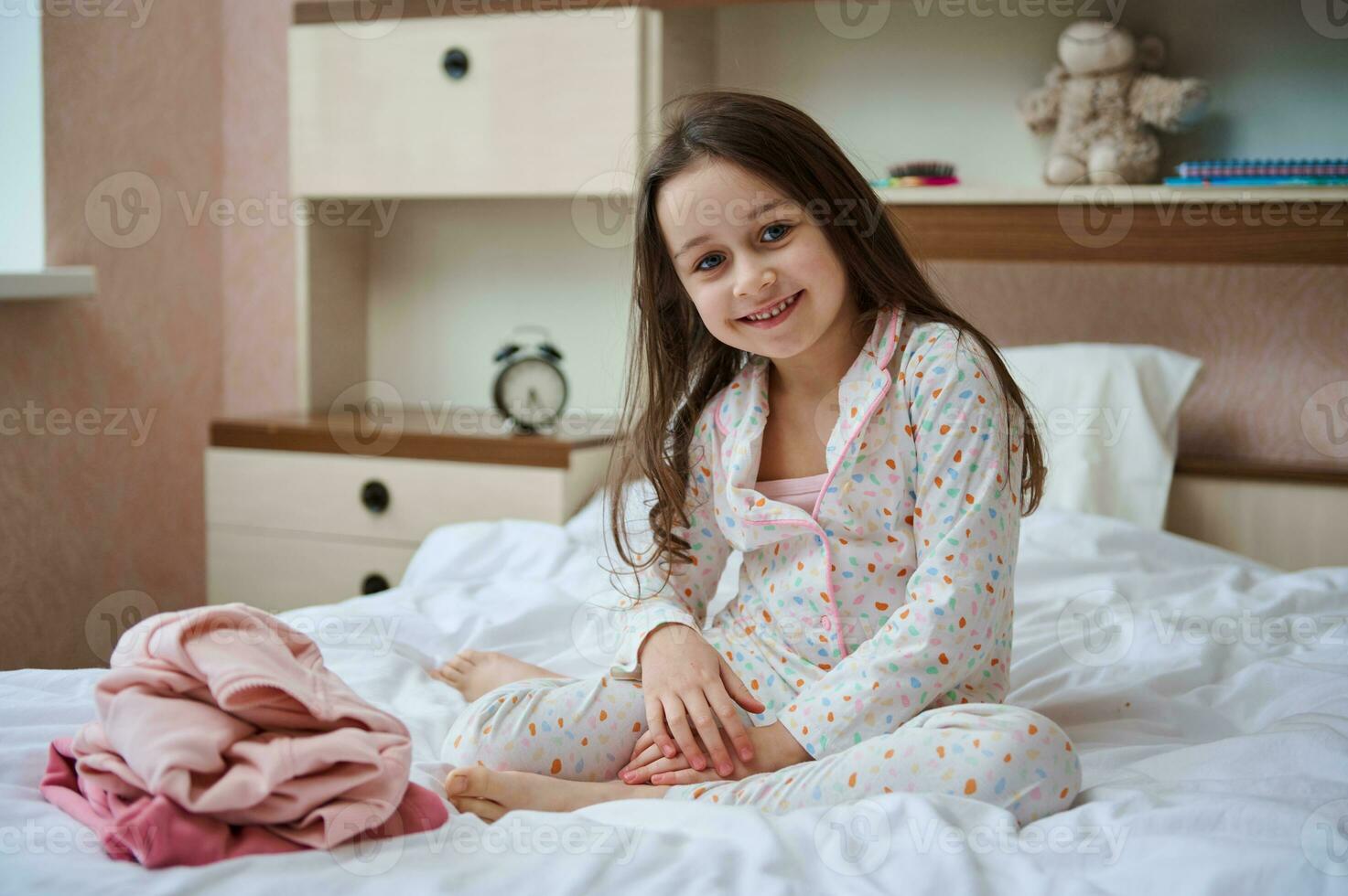 contento poco bambino ragazza nel pigiama seduta su il letto vicino sua piegato Abiti sorridente carino guardare a telecamera foto