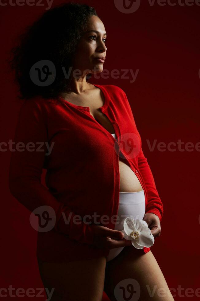 latino americano etnico donna nel rosso camicia, Tenere bianca orchidea fiore al di sopra di sua incinta gonfiarsi, guardare sognante a parte foto