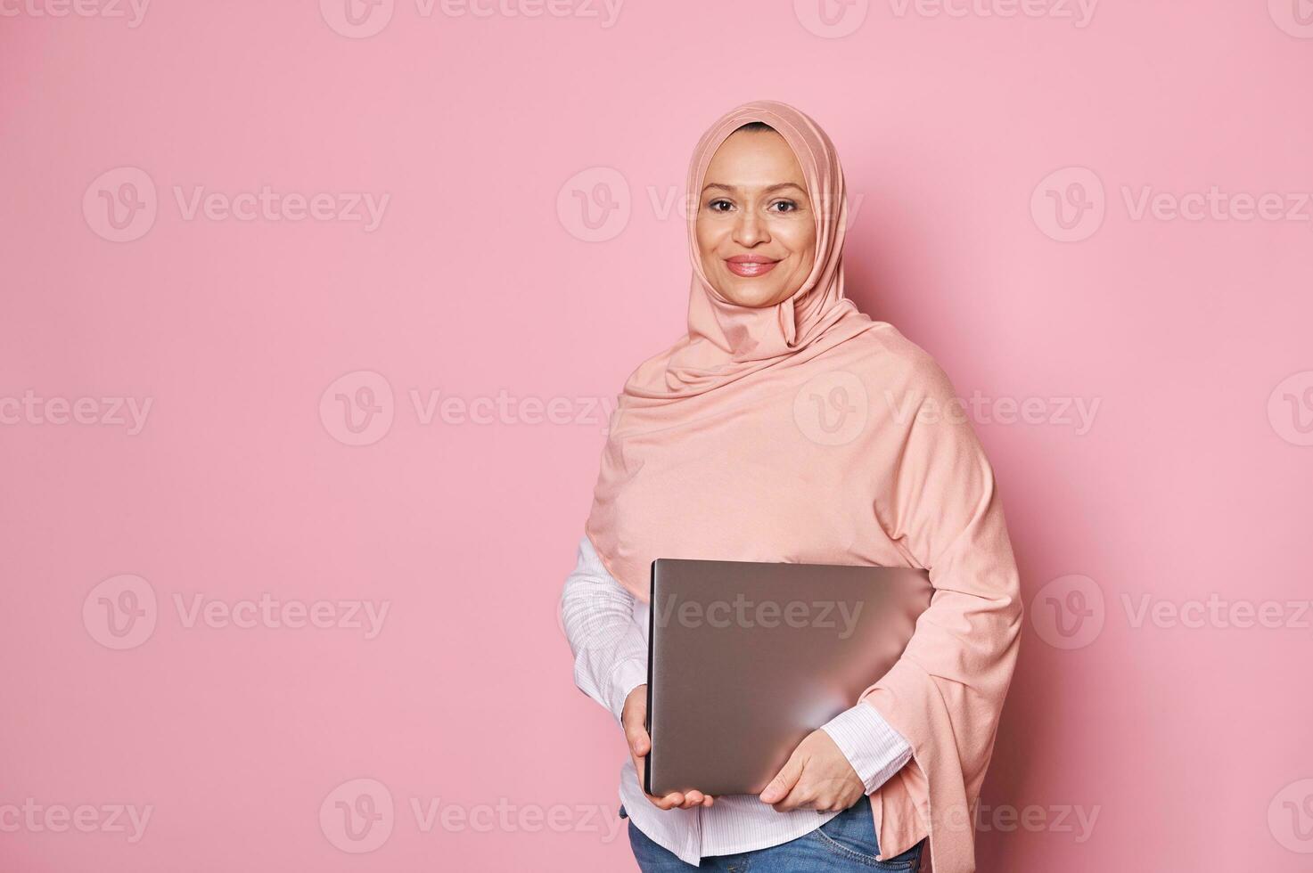 fiducioso arabo musulmano donna nel rosa hijab, Tenere un' computer portatile, sorridente carino guardare a telecamera, isolato rosa sfondo foto