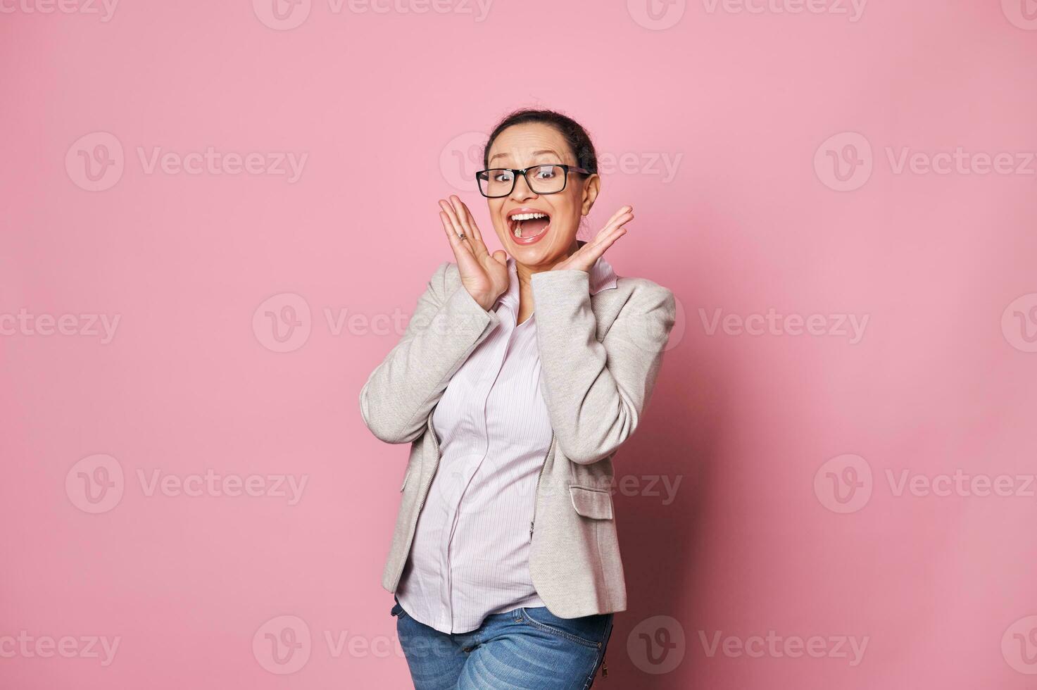 stupito contento bellissimo incinta emotivo donna nel di moda occhiali, in posa con Aperto bocca, isolato al di sopra di rosa sfondo foto