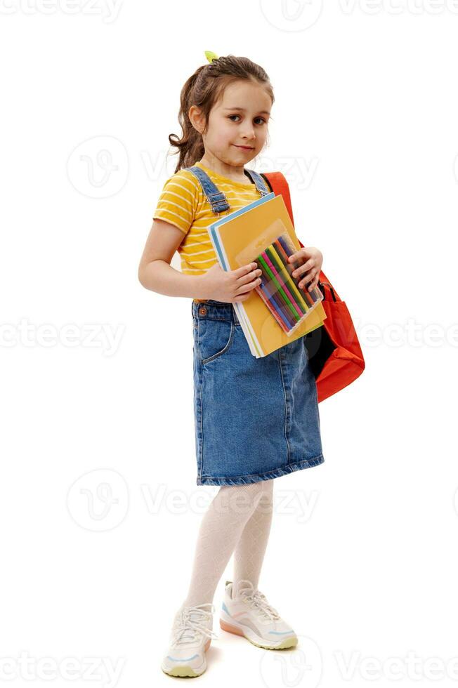 inteligente scolara nel giallo maglietta e denim prendisole, trasporto zaino e scuola forniture, sorrisi su bianca sfondo foto