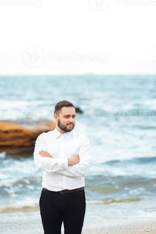 sposo in camicia bianca e pantaloni neri in riva al mare foto