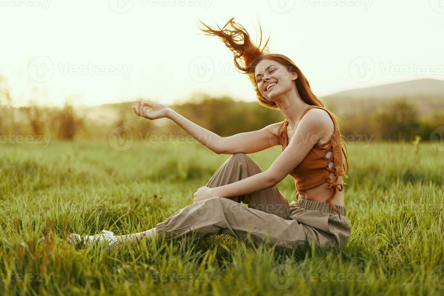 un' donna si siede su il verde giovane fresco erba nel il Aperto aria e ride e sorrisi felicemente nel il tramonto luce, sua volante rosso capelli svolazzanti nel il vento foto