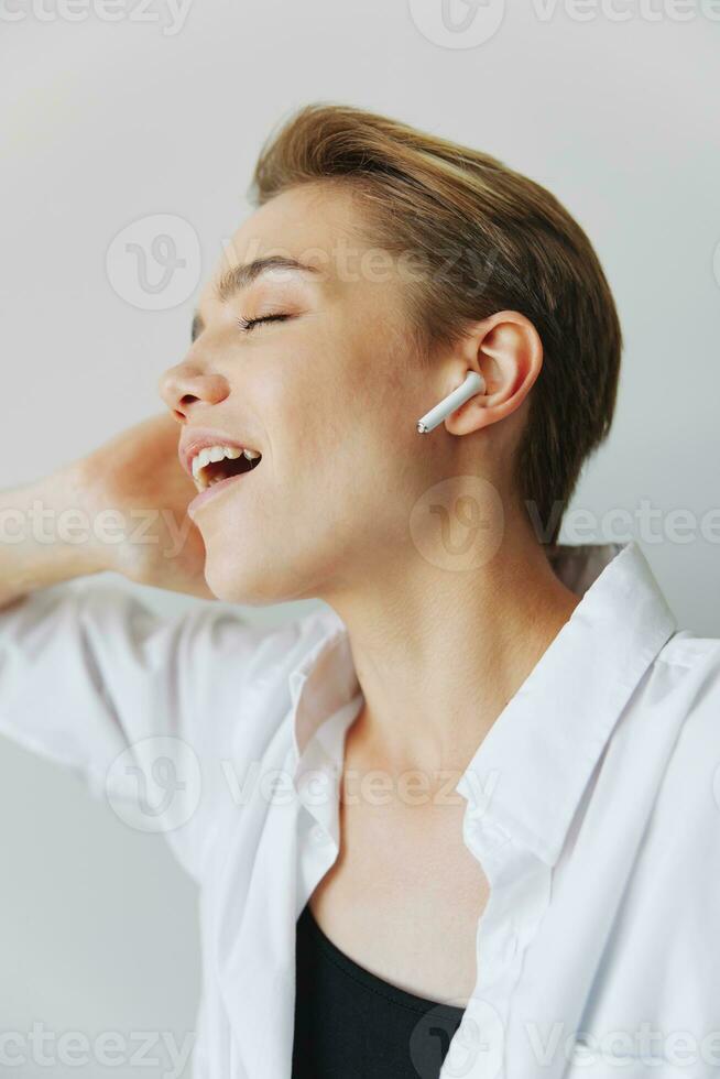 giovane donna adolescente ascoltando per musica con sterile cuffie e danza casa, ghignante con denti con un' corto taglio di capelli nel un' bianca camicia su un' bianca sfondo. ragazza naturale pose con no filtri foto