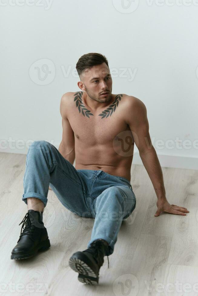 modellazione istantanee. grave attraente nudo bello uomo nel jeans seduta su pavimento in posa nel bianca studio sfondo. moda uomini sparare concetto. copia spazio. freddo offrire per anno Domini foto