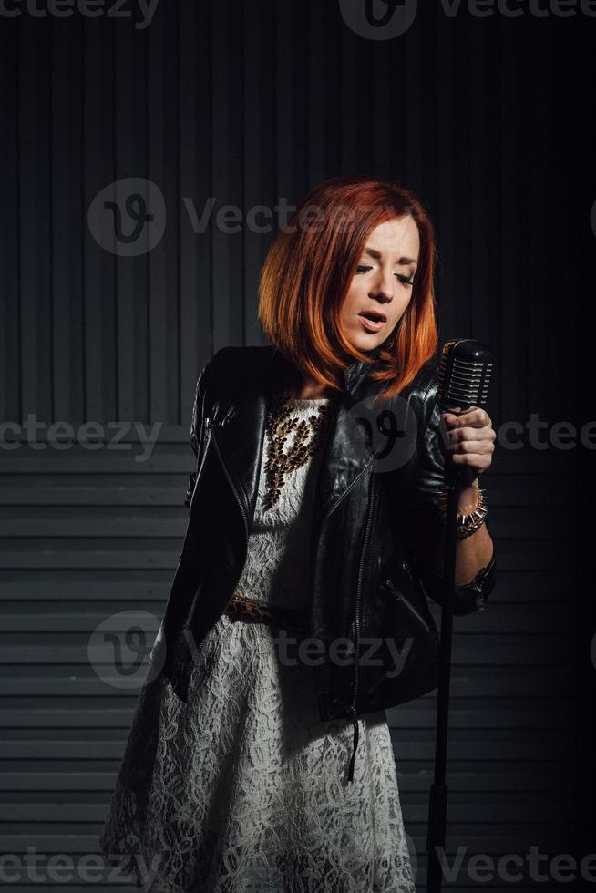 giovane ragazza dai capelli rossi in un abito bianco e una giacca rocker in pelle nera canta nel microfono foto