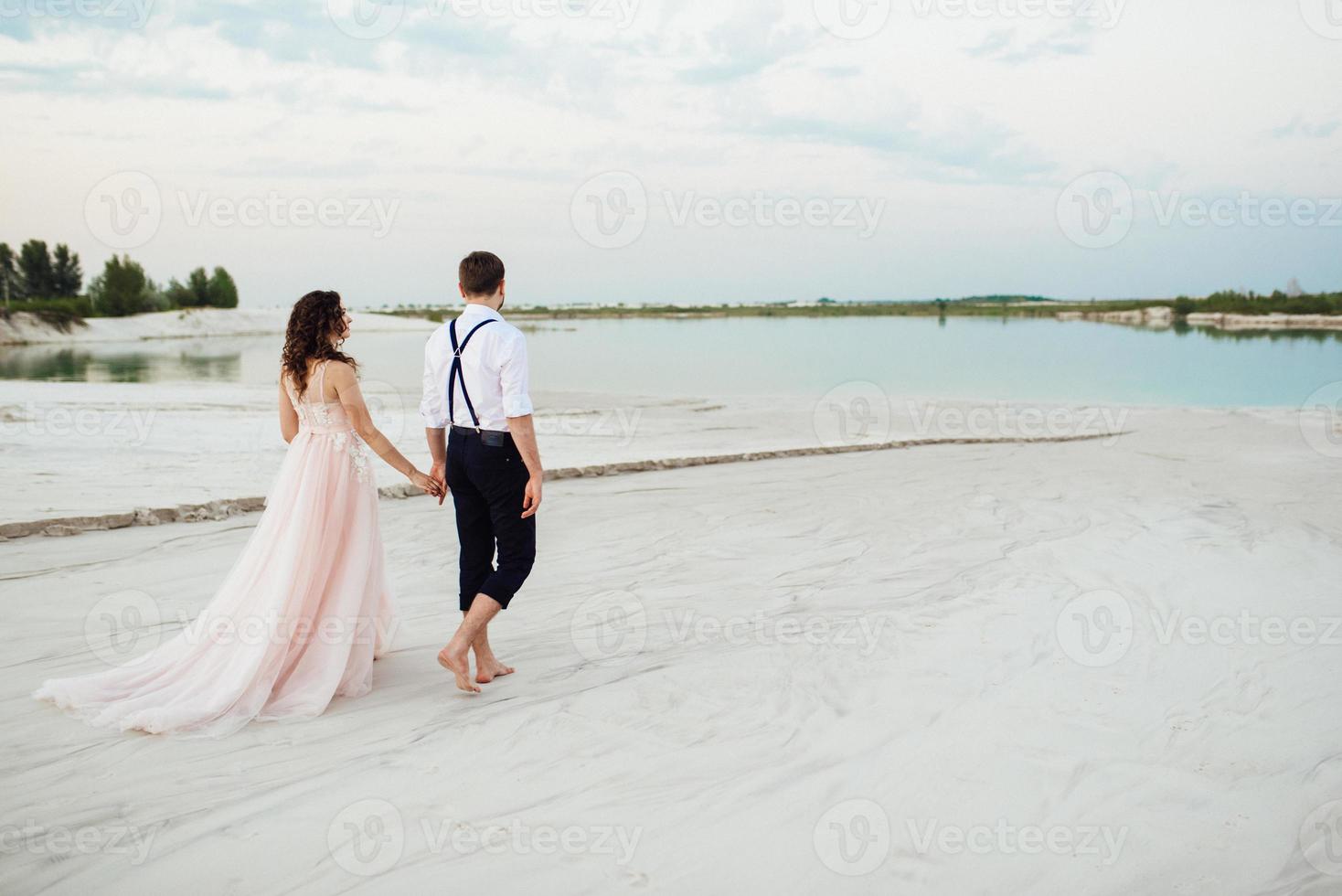 giovane coppia un ragazzo in calzoni neri e una ragazza in un vestito rosa stanno camminando lungo la sabbia bianca foto