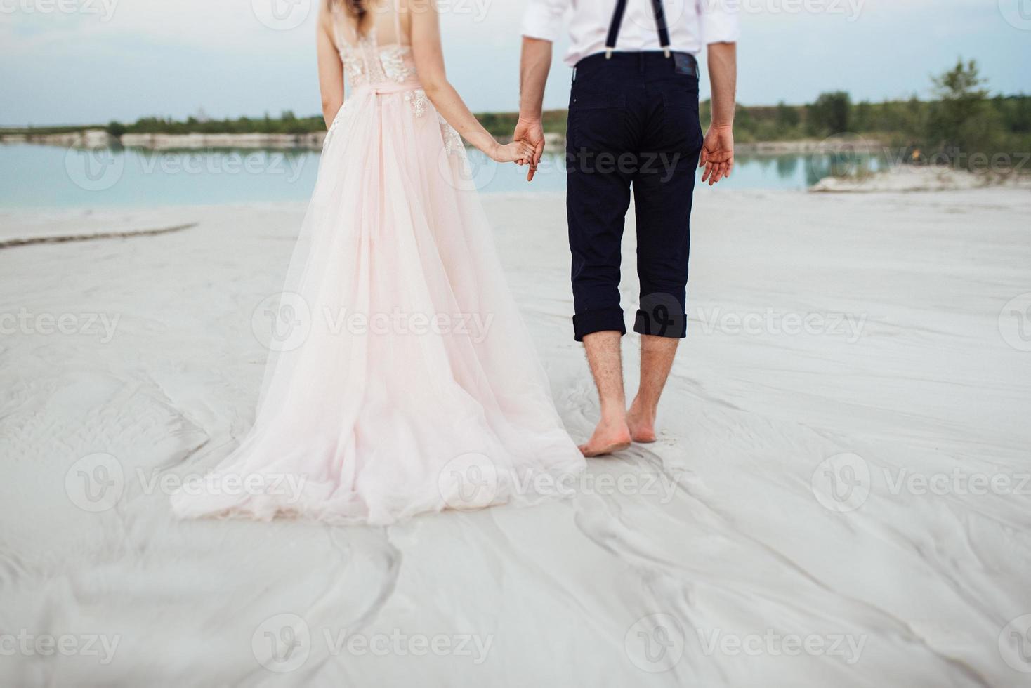 giovane coppia un ragazzo in calzoni neri e una ragazza in un vestito rosa stanno camminando lungo la sabbia bianca foto