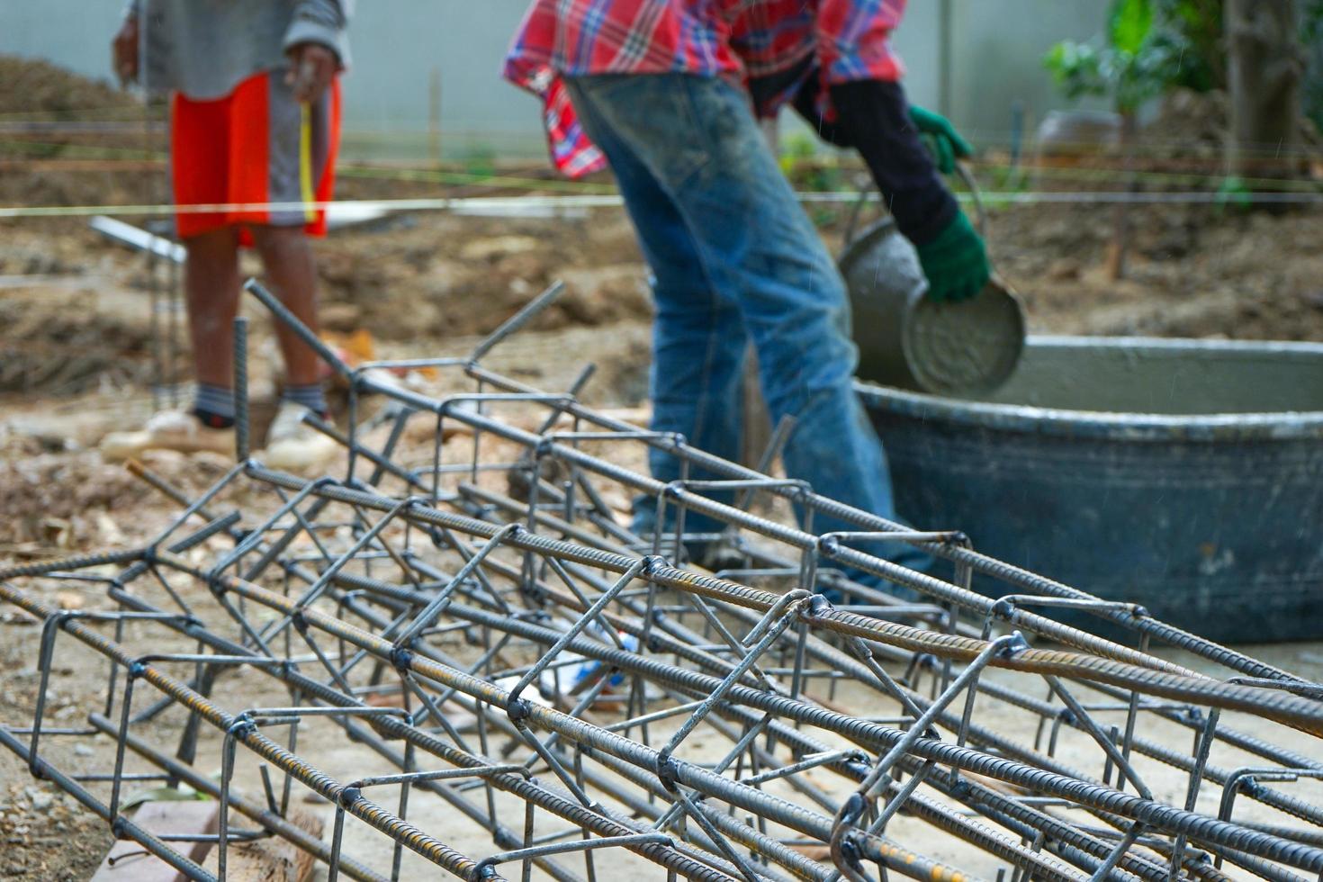 primo piano mucchio di acciaieria di rinforzo sul terreno in cantiere con lavoratori offuscati che trasportano il secchio di cemento misto foto