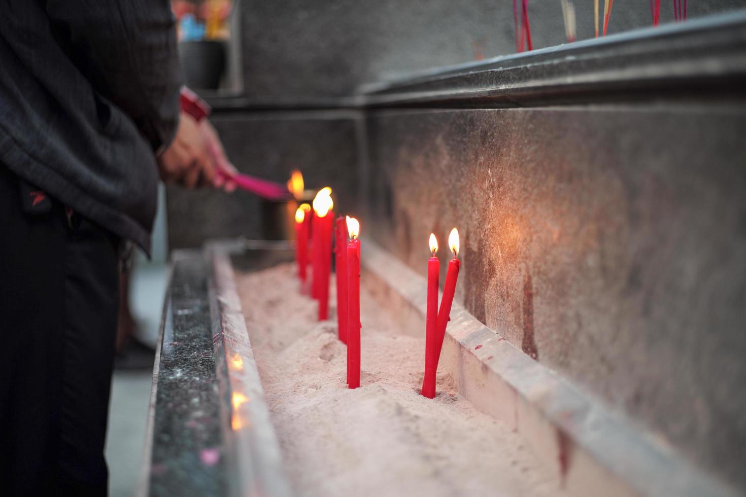 gruppo di candele di illuminazione rosse messe nella sabbia in un vassoio di cemento con mani sfocate che tengono incensi foto