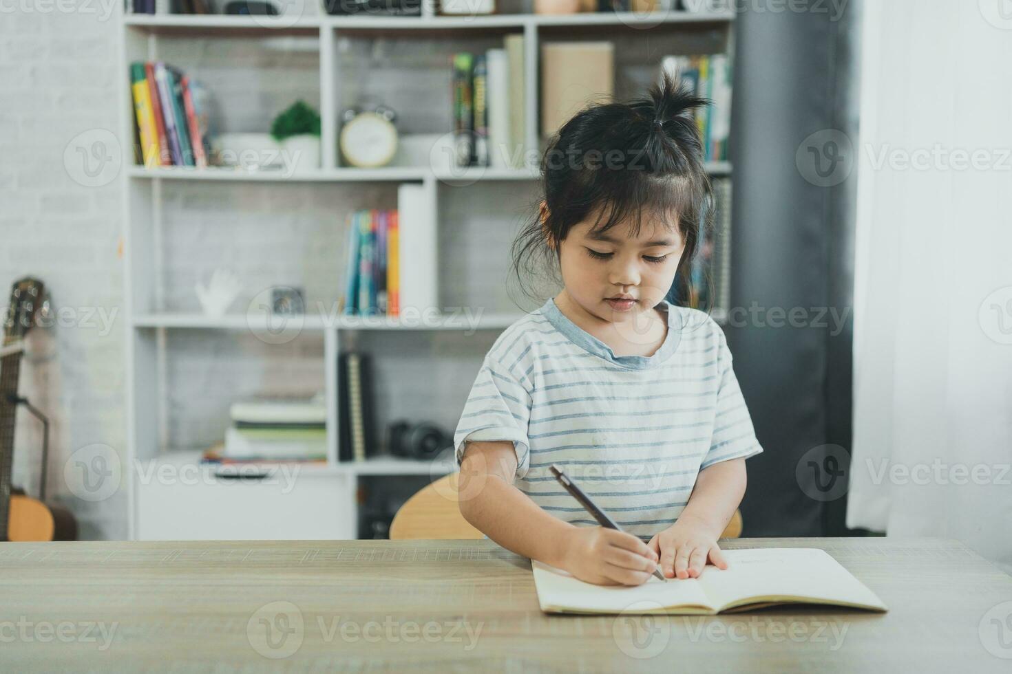 asiatico bambino ragazza indossare un' blu a strisce camicia Scrivi Appunti nel taccuino per studia in linea su legna tavolo scrivania nel vivente camera a casa. formazione scolastica apprendimento in linea a partire dal casa concetto. foto