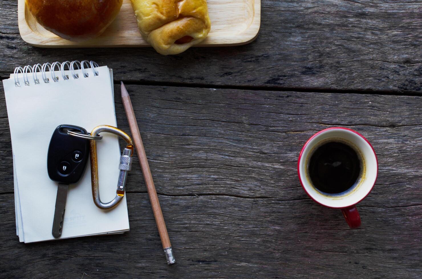 chiavi della macchina sul blocco note con matita e tazza di caffè rosso e pane panino sul fondo della tavola in legno foto