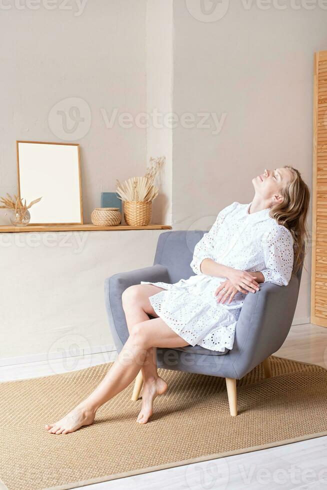 bellissimo donna nel moda vestito seduta su morbido sedia a piedi nudi su bianca parete sfondo foto
