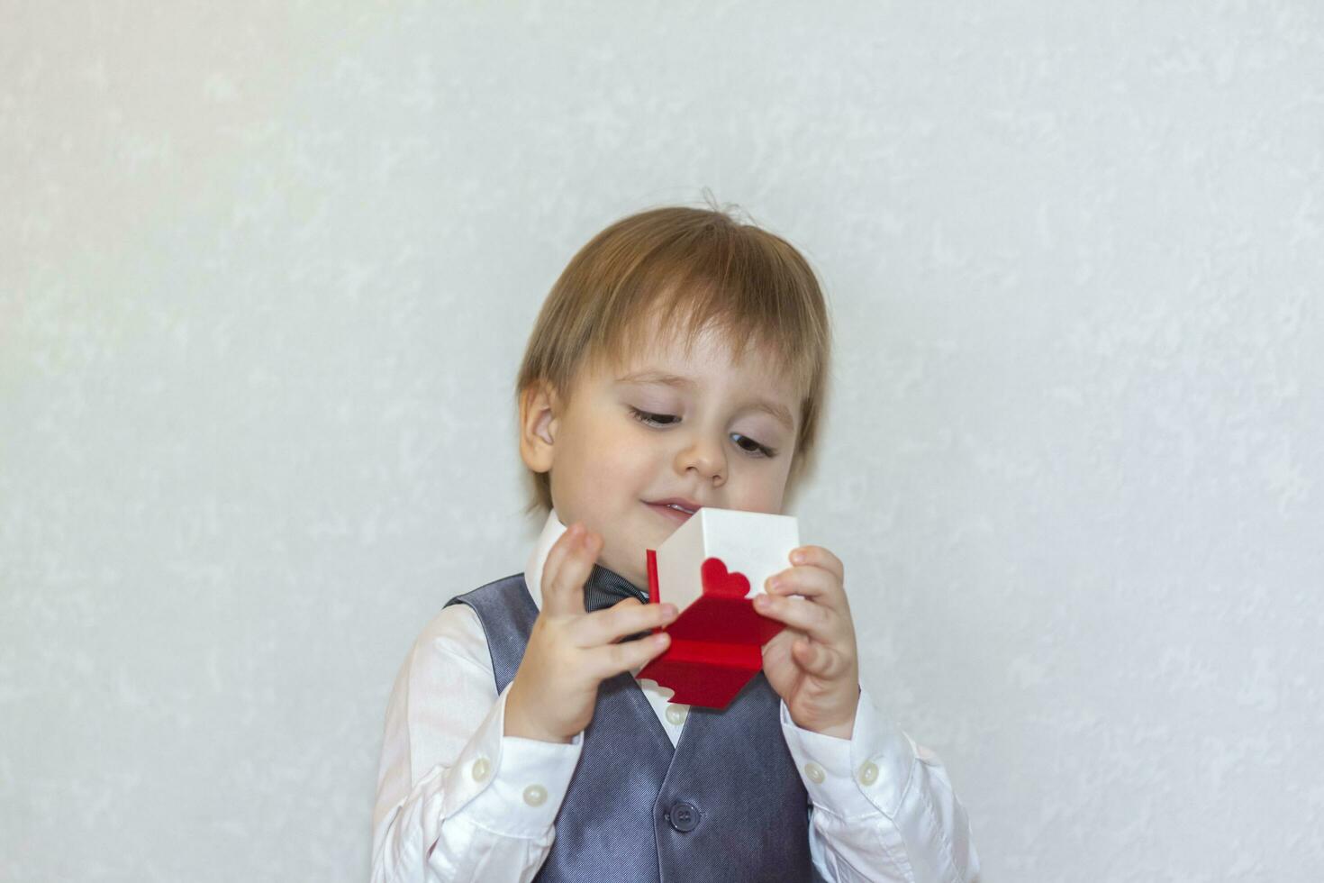 un' poco ragazzo detiene e mani al di sopra di un' rosso scatola, un' San Valentino giorno tema concetto. ritratto di un' carino ragazzo nel un' completo da uomo con un' arco legare. foto