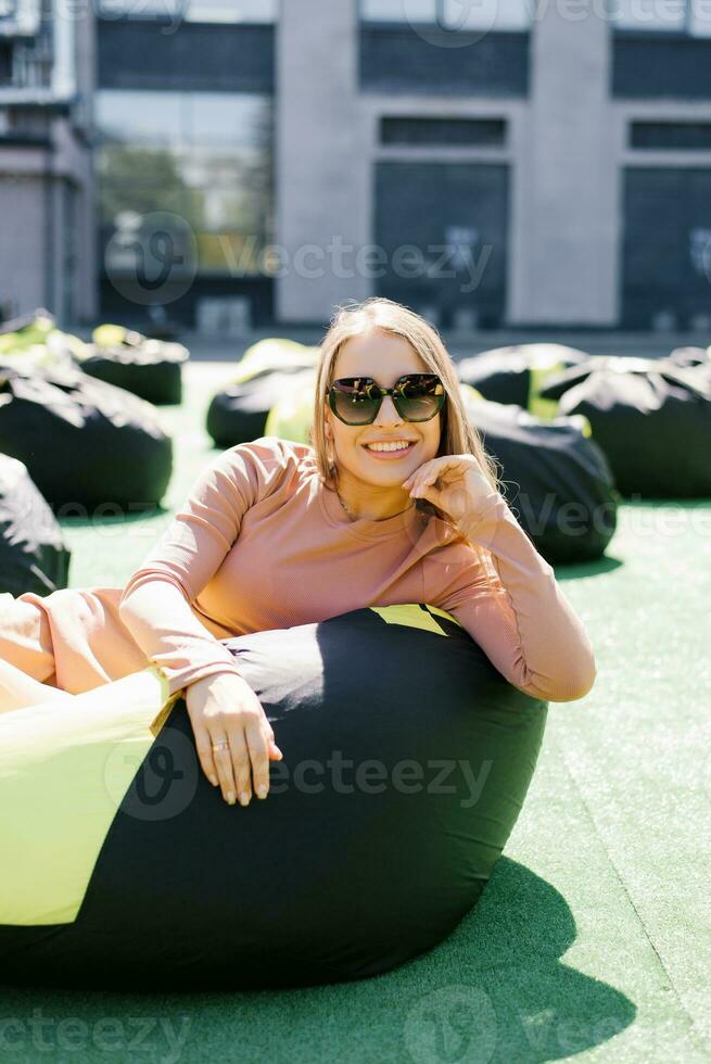 giovane donna è seduta su un facile sedia all'aperto e godendo un' soleggiato giorno. un' bellissimo contento ragazza nel occhiali da sole. foto
