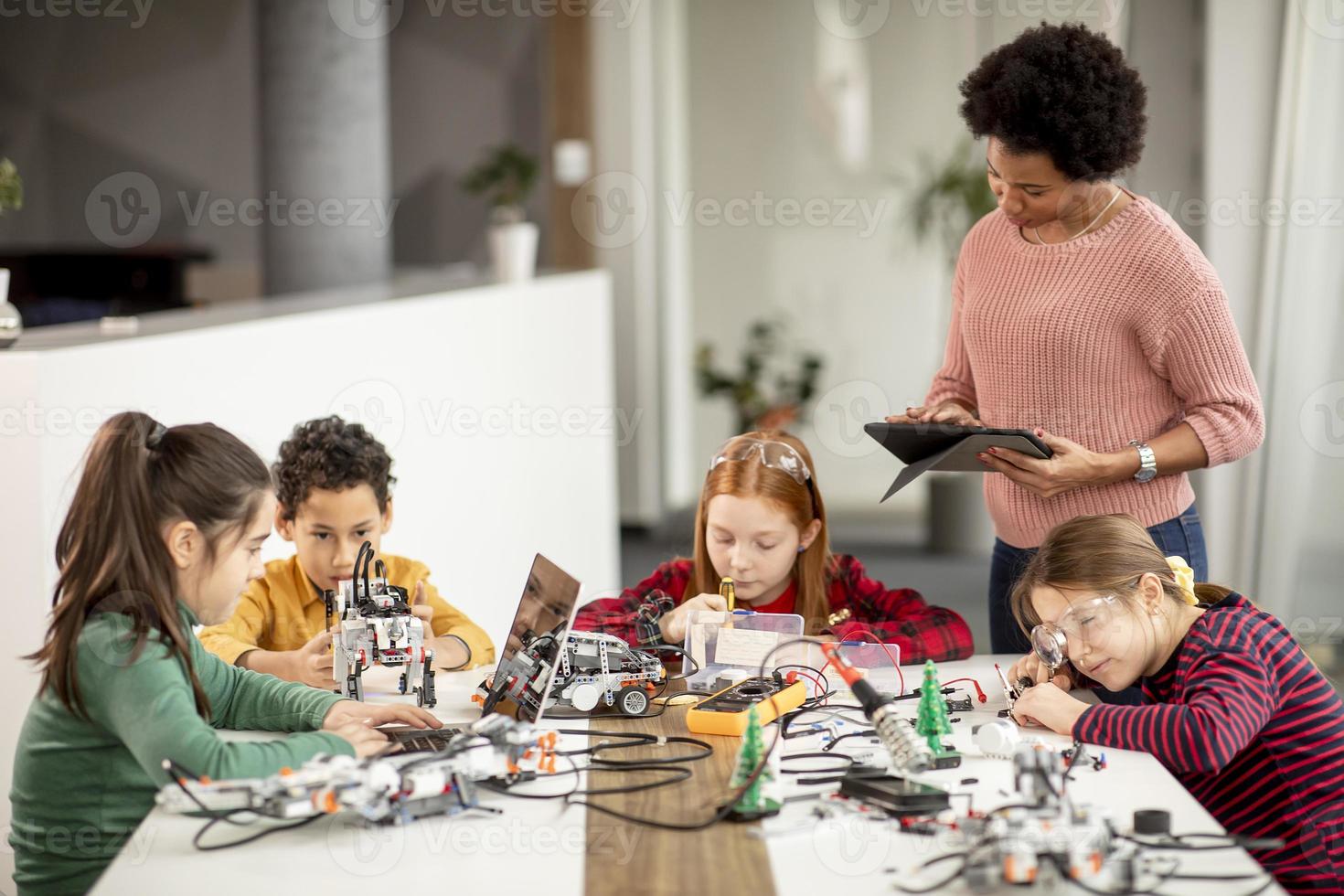 bambini felici con la loro insegnante di scienze afroamericana con laptop che programmano giocattoli elettrici e robot in classe di robotica foto