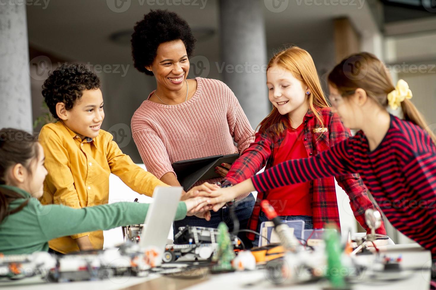 bambini felici con la loro insegnante di scienze afroamericana con laptop che programmano giocattoli elettrici e robot in classe di robotica foto