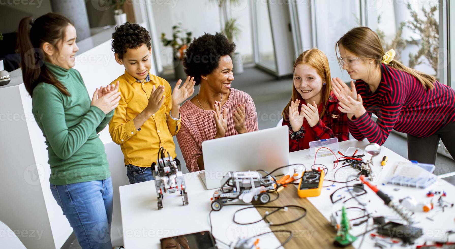 bambini felici con la loro insegnante di scienze afroamericana con laptop che programmano giocattoli elettrici e robot in classe di robotica foto