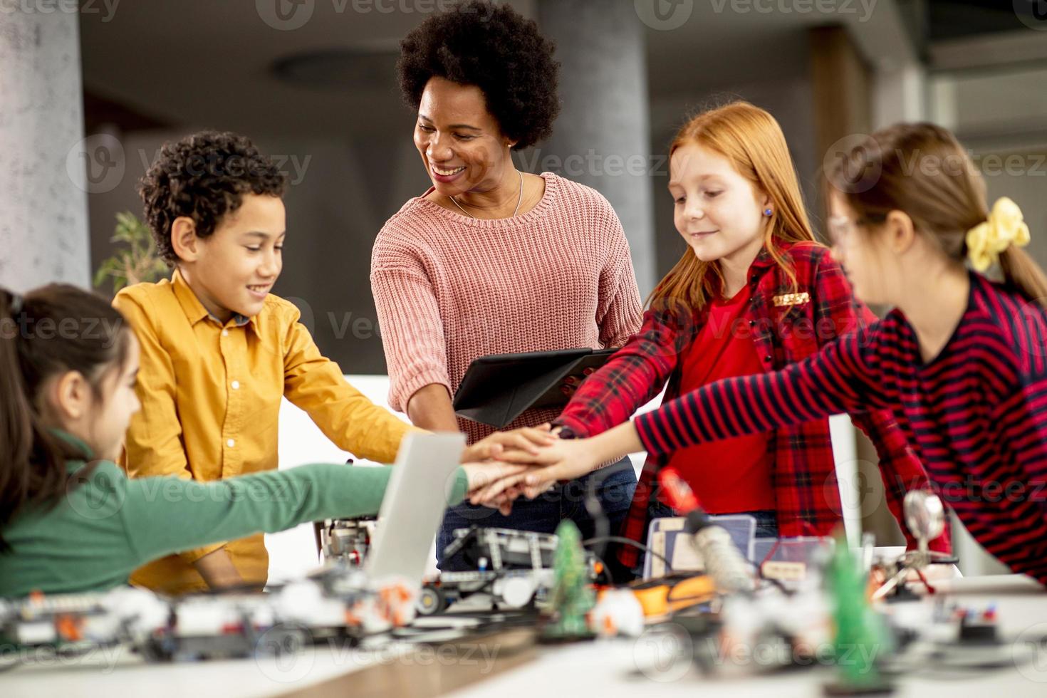 bambini felici con la loro insegnante di scienze afroamericana con laptop che programmano giocattoli elettrici e robot in classe di robotica foto