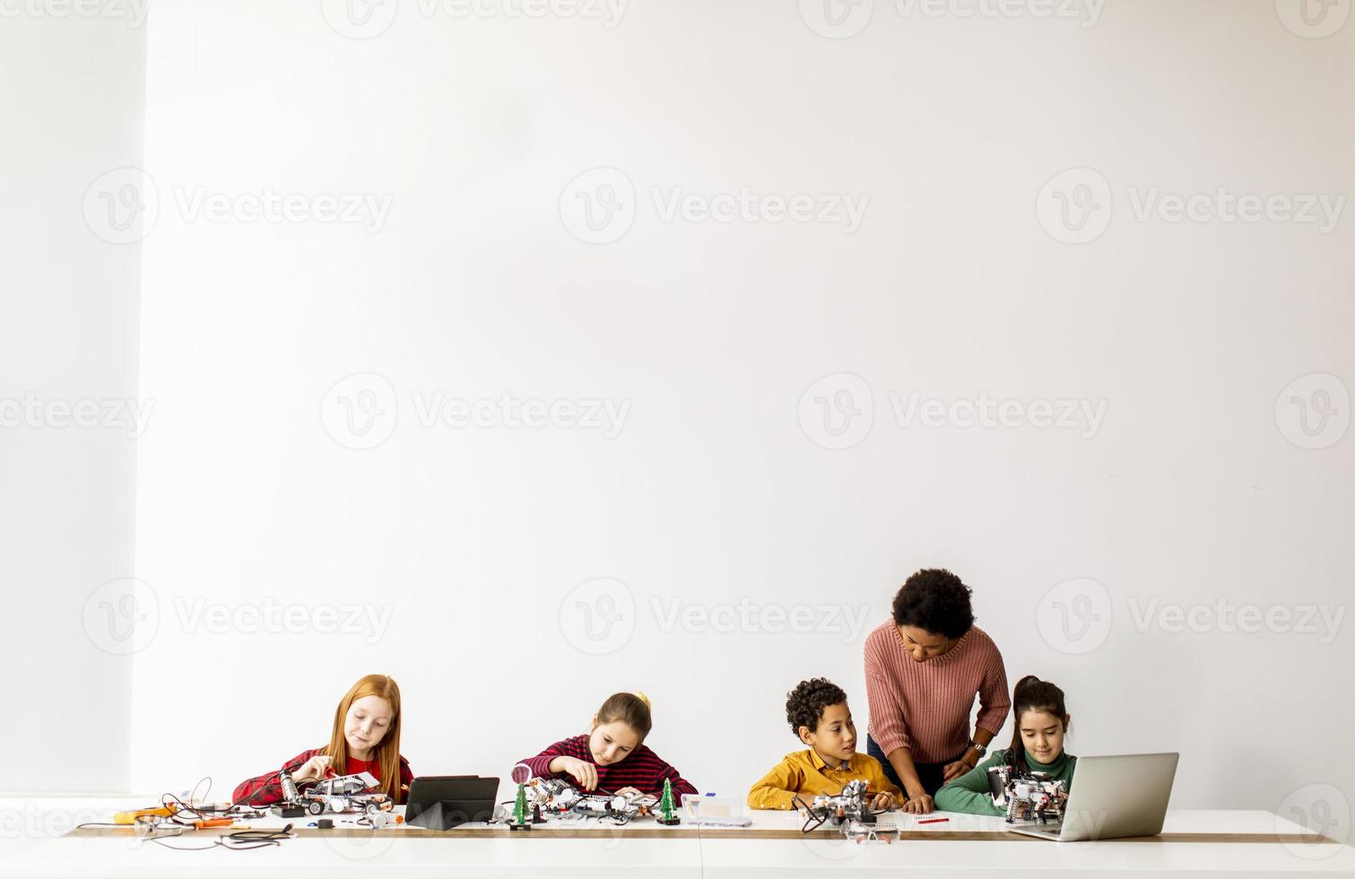 bambini felici con la loro insegnante di scienze afroamericana con laptop che programmano giocattoli elettrici e robot in classe di robotica foto