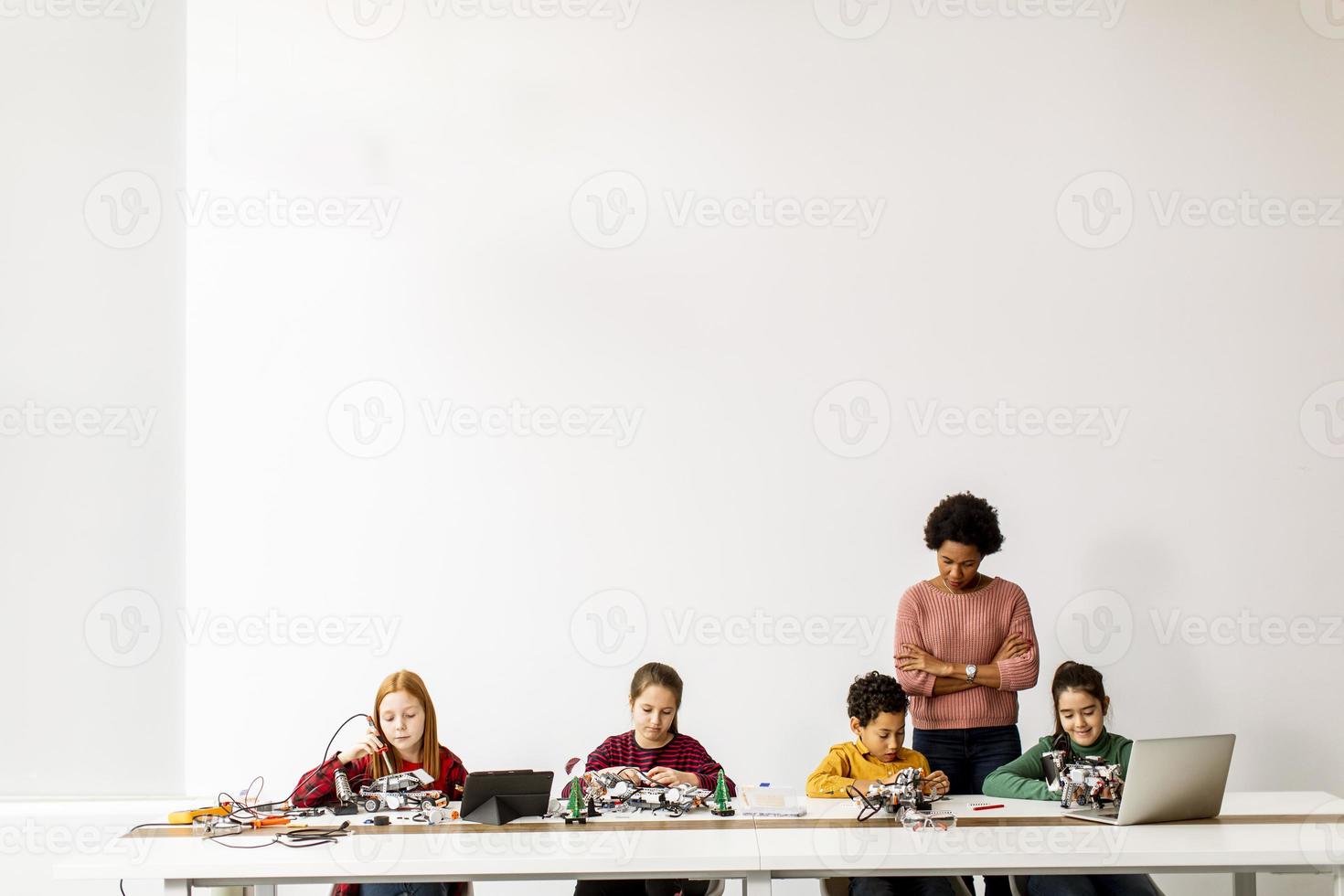 bambini felici con la loro insegnante di scienze afroamericana con laptop che programmano giocattoli elettrici e robot in classe di robotica foto