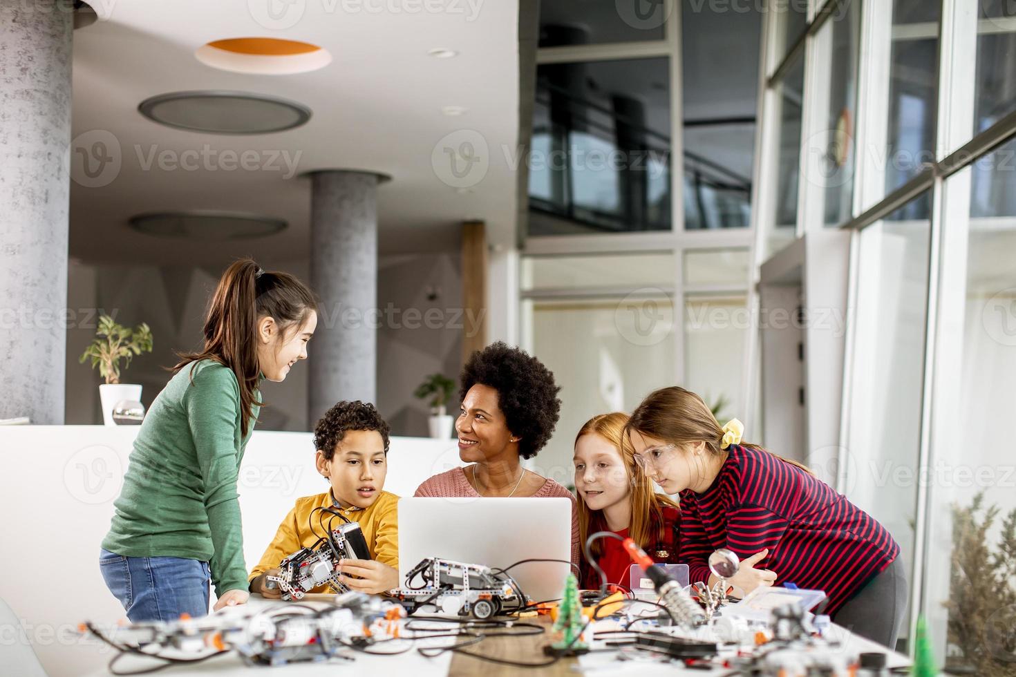 bambini felici con la loro insegnante di scienze afroamericana con laptop che programmano giocattoli elettrici e robot in classe di robotica foto