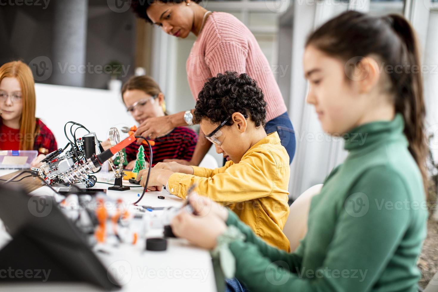 insegnante di scienze femminile afroamericana con un gruppo di bambini che programmano giocattoli elettrici e robot in aula di robotica foto