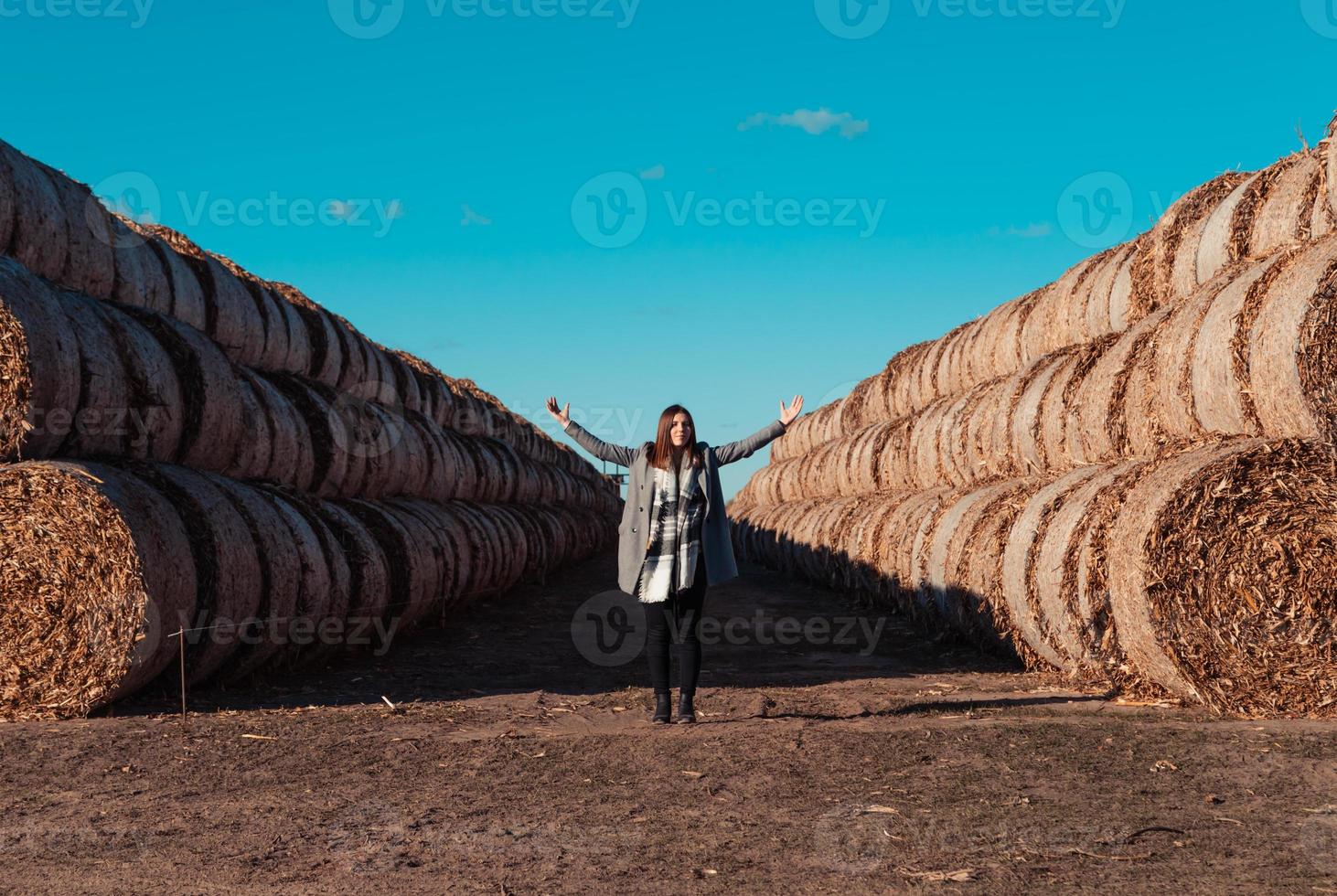 una ragazza di aspetto europeo con un cappotto grigio si trova in un campo vicino a una balla più grande con fieno foto