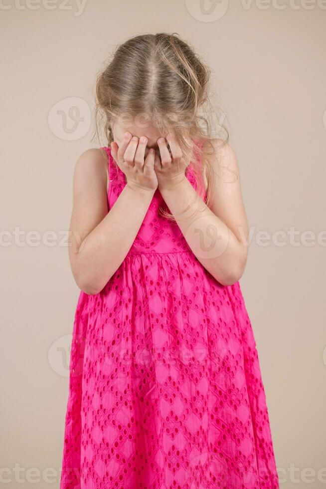 poco ragazza nel un' rosa vestito su un' beige sfondo coperture sua viso con sua mani foto