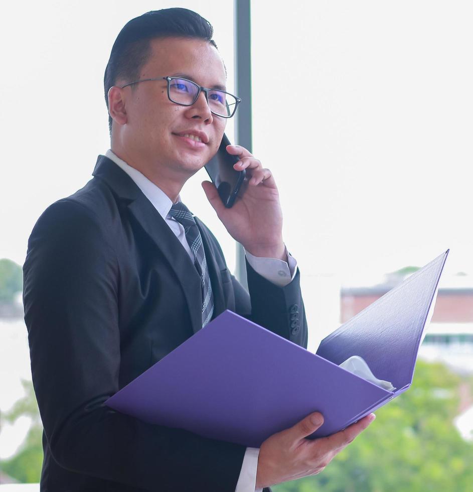 asiatico bel giovane uomo d'affari in possesso di un file di documento per controllare le prestazioni dell'azienda e utilizzando uno smartphone nell'ufficio moderno foto
