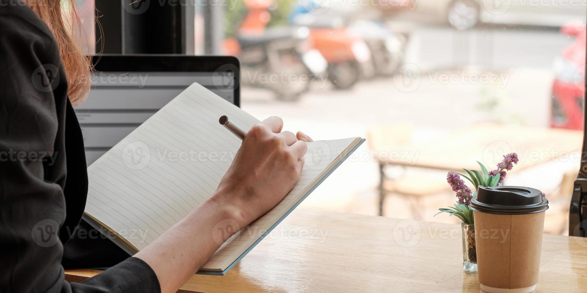 primo piano di donna studio in linea distante su laptop prendere nota scrivere in taccuino seduto sulla scrivania a casa smart donna scrittore o studente scrivere a mano in cancelleria pianificazione blocco note foto