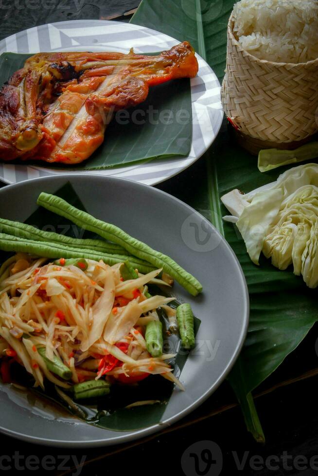 tailandese cibo piatto tutti e due nel Tailandia e Asia, papaia insalata o come noi chiamata esso somtum è integrato con grigliato pollo e appiccicoso riso con fresco patatine fritte. servito su il nero di legno tavolo. foto