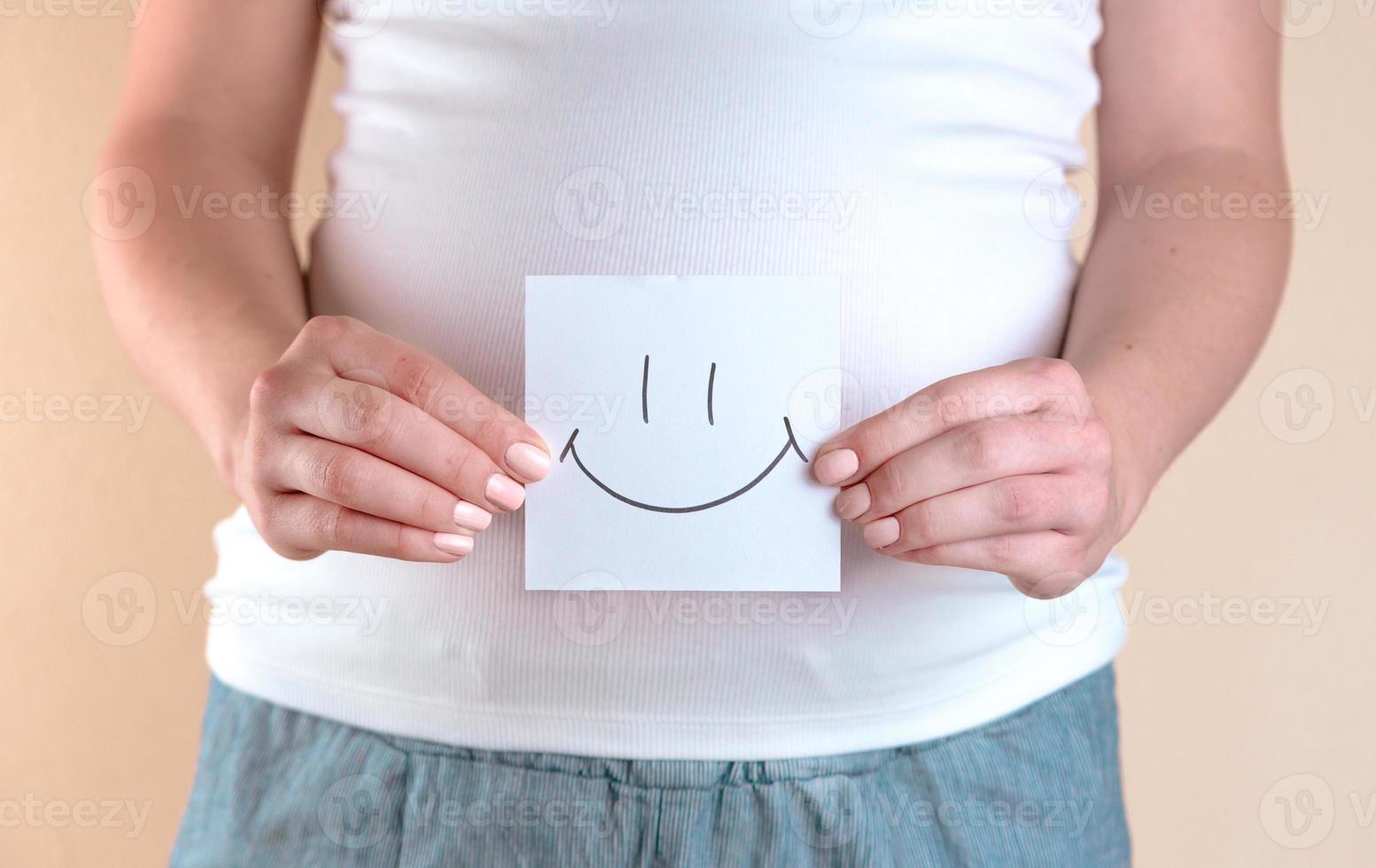 una vista ravvicinata della pancia di una donna incinta che tiene un pezzo di carta con una faccina sorridente foto