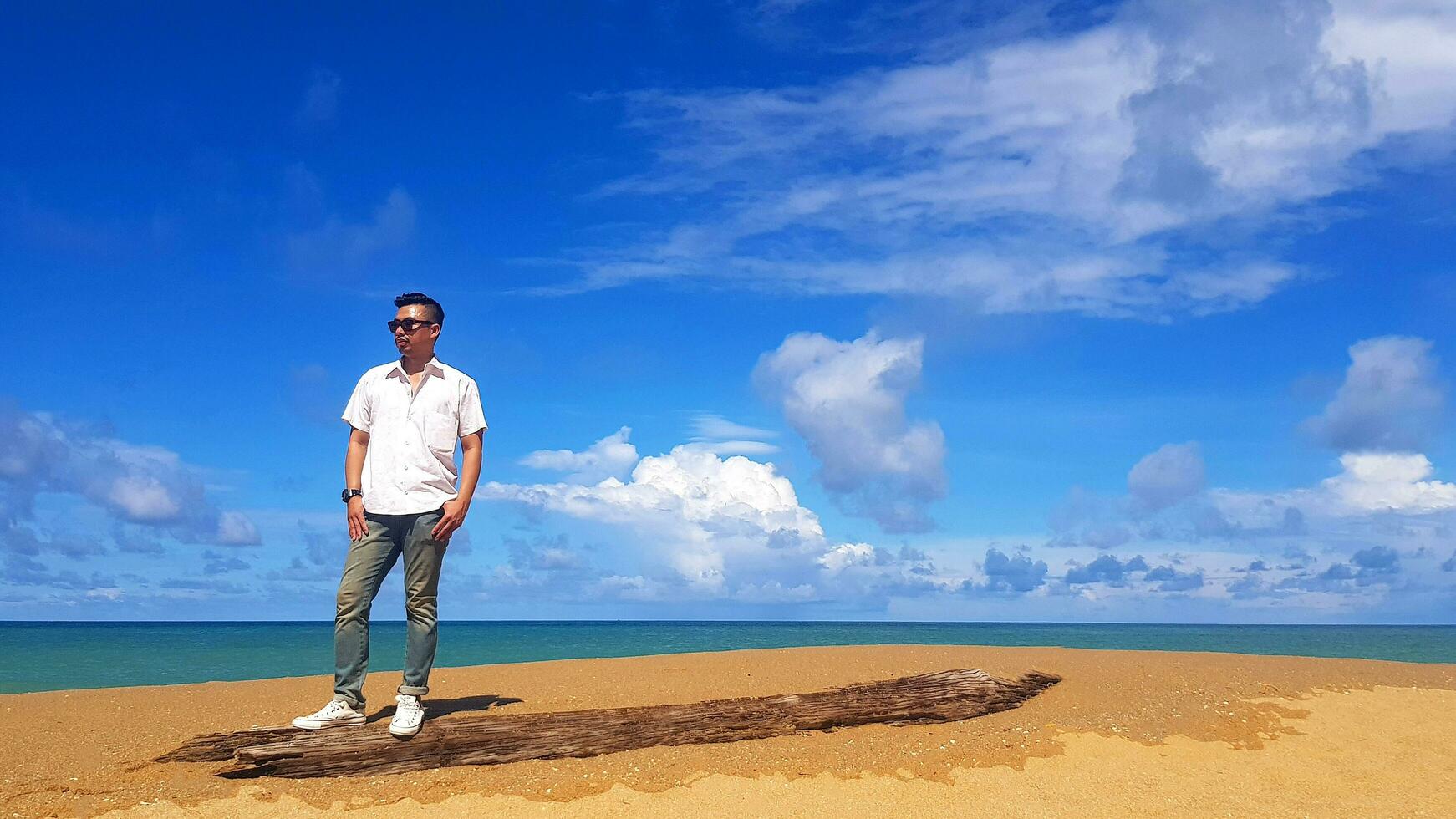 asiatico uomo nel bianca camicia e jeans, indossare occhiali da sole, In piedi su legname con mare, blu cielo e nube sfondo con copia spazio a Mai khao spiaggia Phuket, Tailandia. turista, relax, punto di riferimento, viaggio. foto
