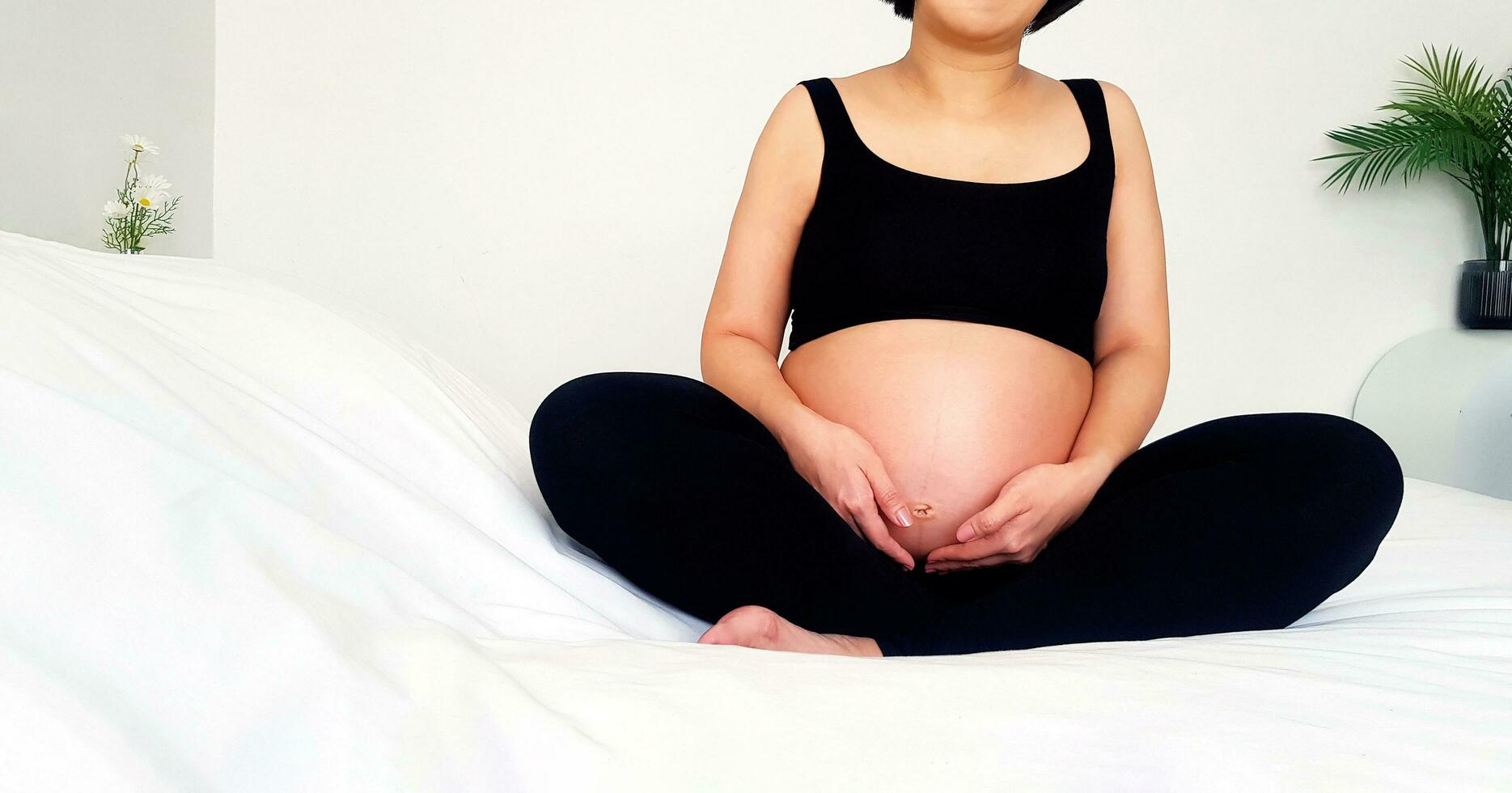 asiatico incinta donna nel nero maglietta seduta su bianca letto e toccante sua pancia nel il Camera da letto con copia spazio. gravidanza 7-8 mesi, maternità, amore, aspettativa, cura e in attesa per sua bambino foto