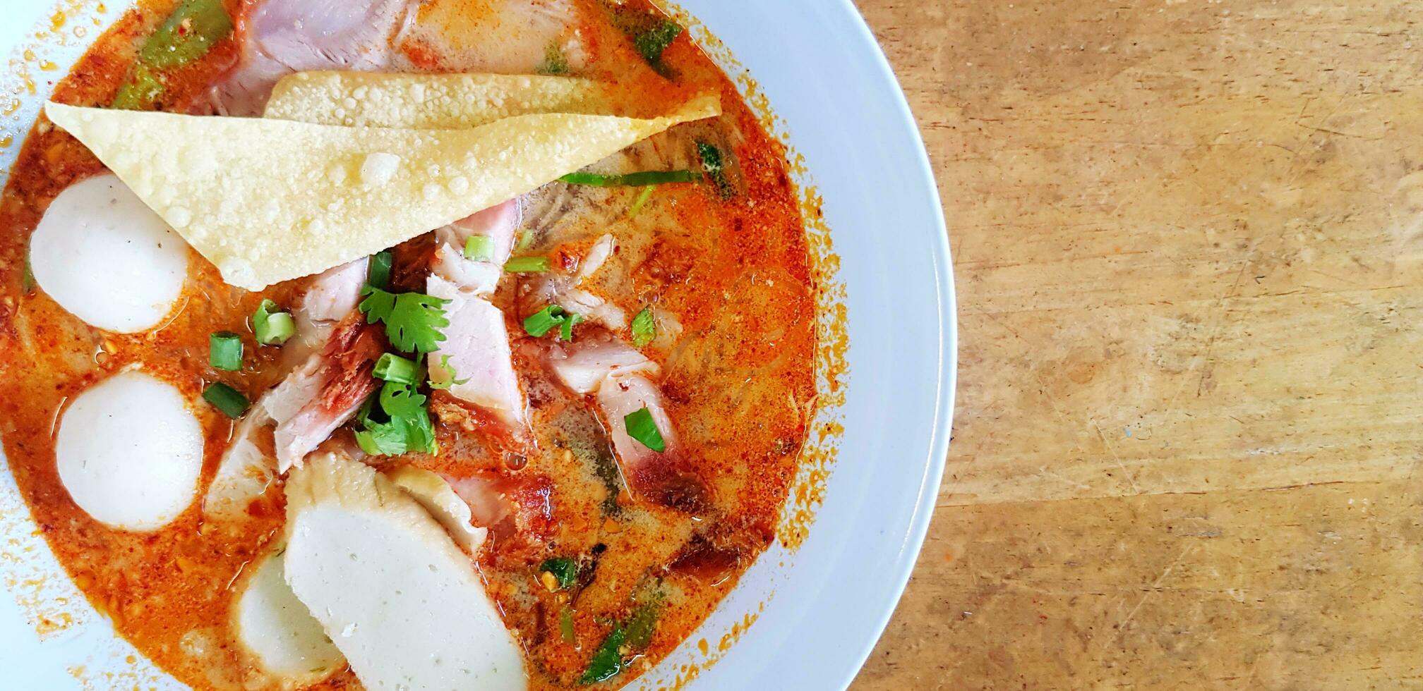 superiore Visualizza caldo speziato spaghetto con la minestra, in profondità fritte won-ton carne, pesce o pollo sfera, croccante Maiale e nel leggero blu ciotola su di legno tavolo o sfondo con sinistra copia spazio. asiatico cibo foto