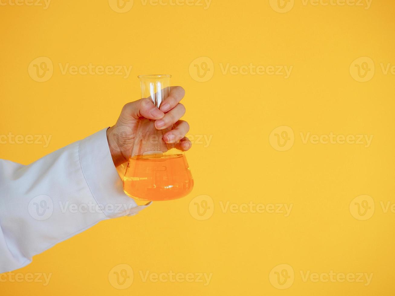 scienziato in abito bianco che tiene un bicchiere di soluzione chimica arancione su sfondo giallo foto