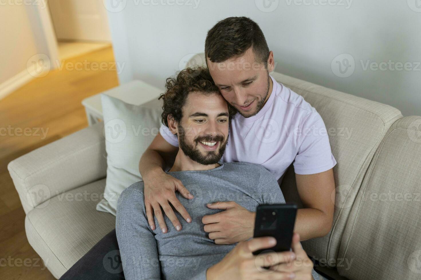 carino giovane gay coppia video chiamata loro amici nel loro vivente camera a casa. Due maschio Gli amanti sorridente allegramente mentre saluto loro amici su un' smartphone. giovane gay coppia seduta insieme. foto