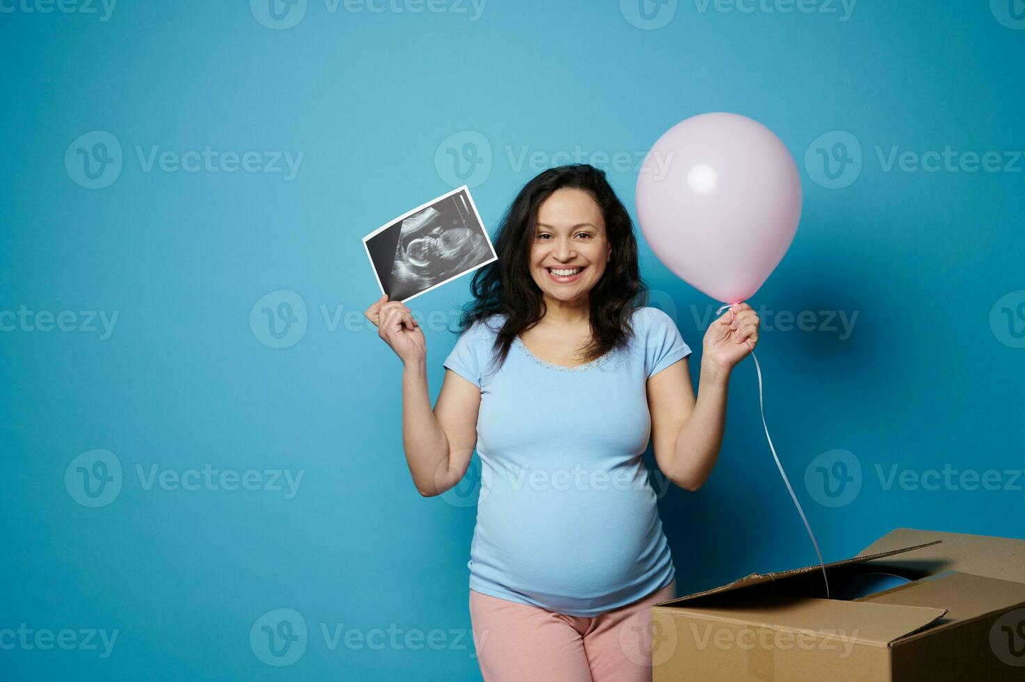 eccitato contento incinta donna sorrisi a telecamera, detiene ultrasuono scansione sua futuro bambino e rosa Palloncino, blu sfondo foto