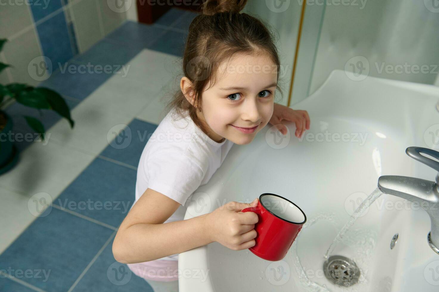 malizioso bambino ragazza in piedi di Lavello nel casa bagno, sorridente guardare a telecamera mentre pulizia bocca, spazzolatura denti foto