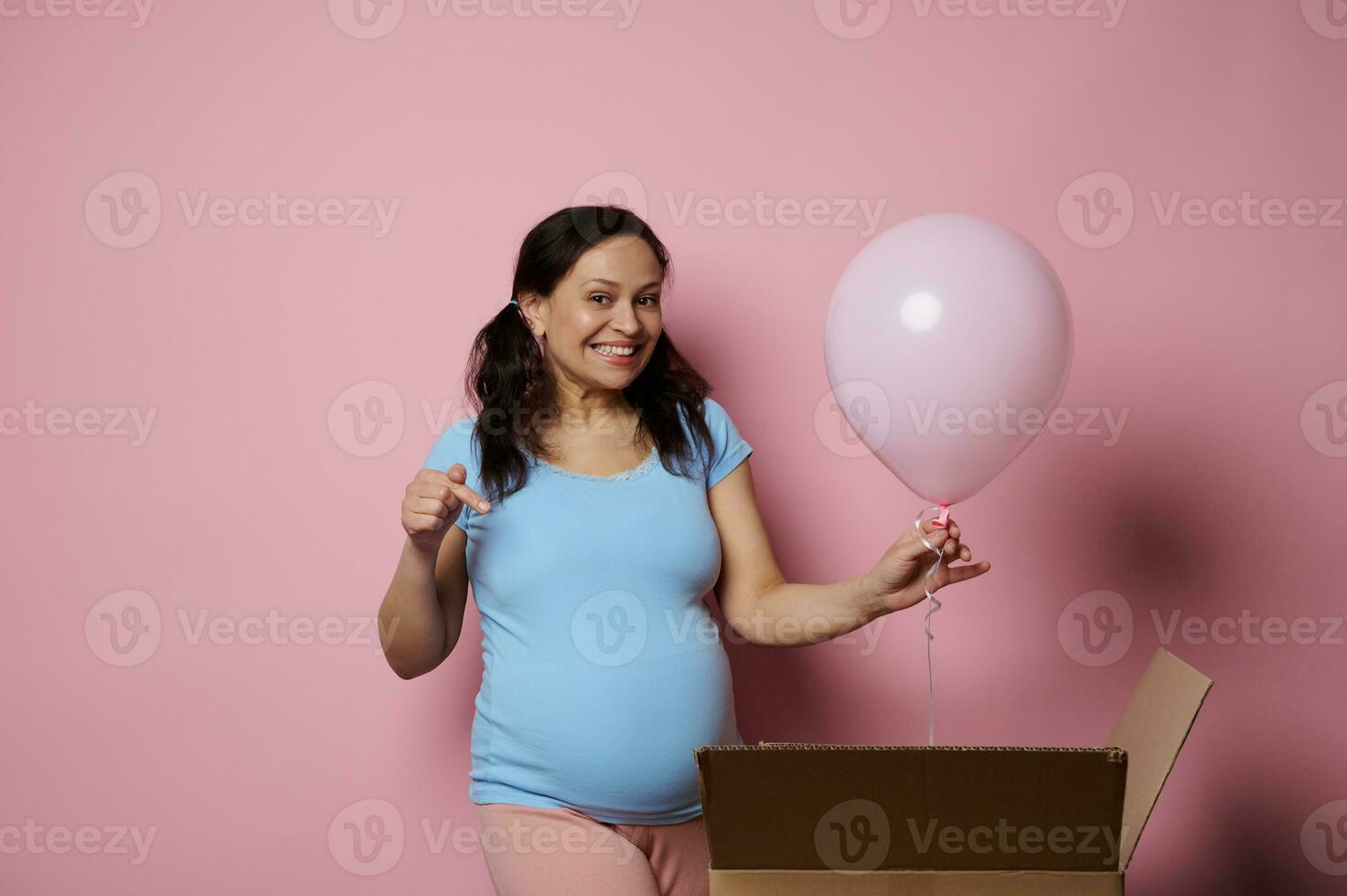 contento incinta donna nel blu maglietta, detiene un' rosa aria Palloncino, punti dito a sua gonfiarsi, contento aspettandosi un' bambino ragazza. foto