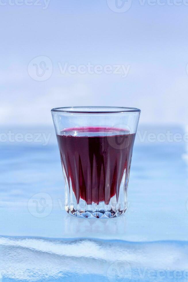 rosso tintura nel un' piccolo bicchiere è su il Ghiaccio. alcool nel un' bicchiere su trasparente Ghiaccio. leggero sfondo sfocato. verticale. foto